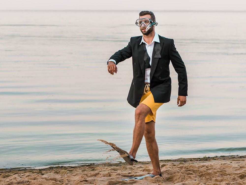 A person walks along a beach wearing flippers, a mask and snorkel, bathing suit and tuxedo jacket and white shirt.