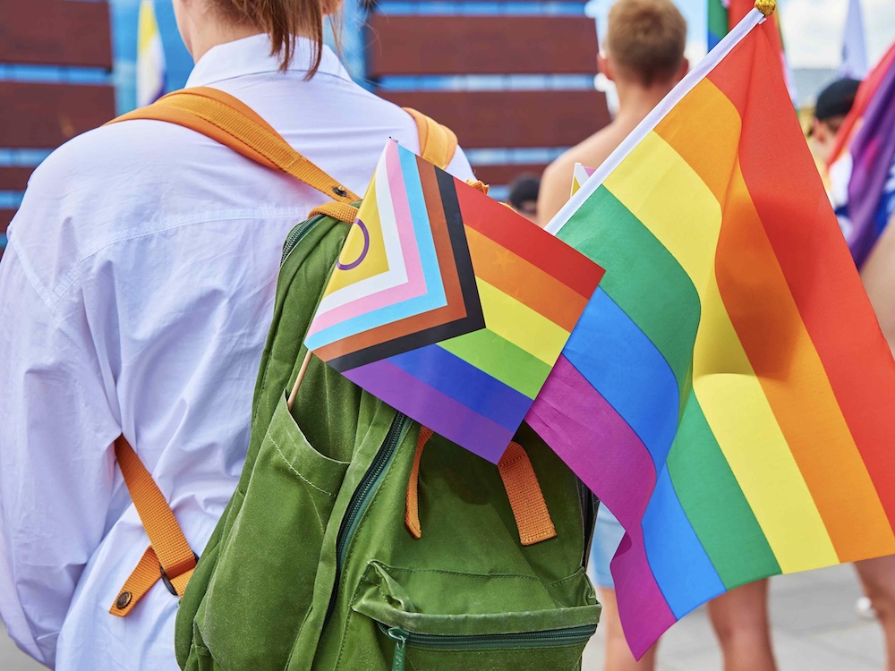 A person is photographed from the back. They are wearing a light blue button-down shirt is wearing a green backpack stuck with two Pride flags. To the right of the frame, other people are carrying Pride flags in an urban environment.