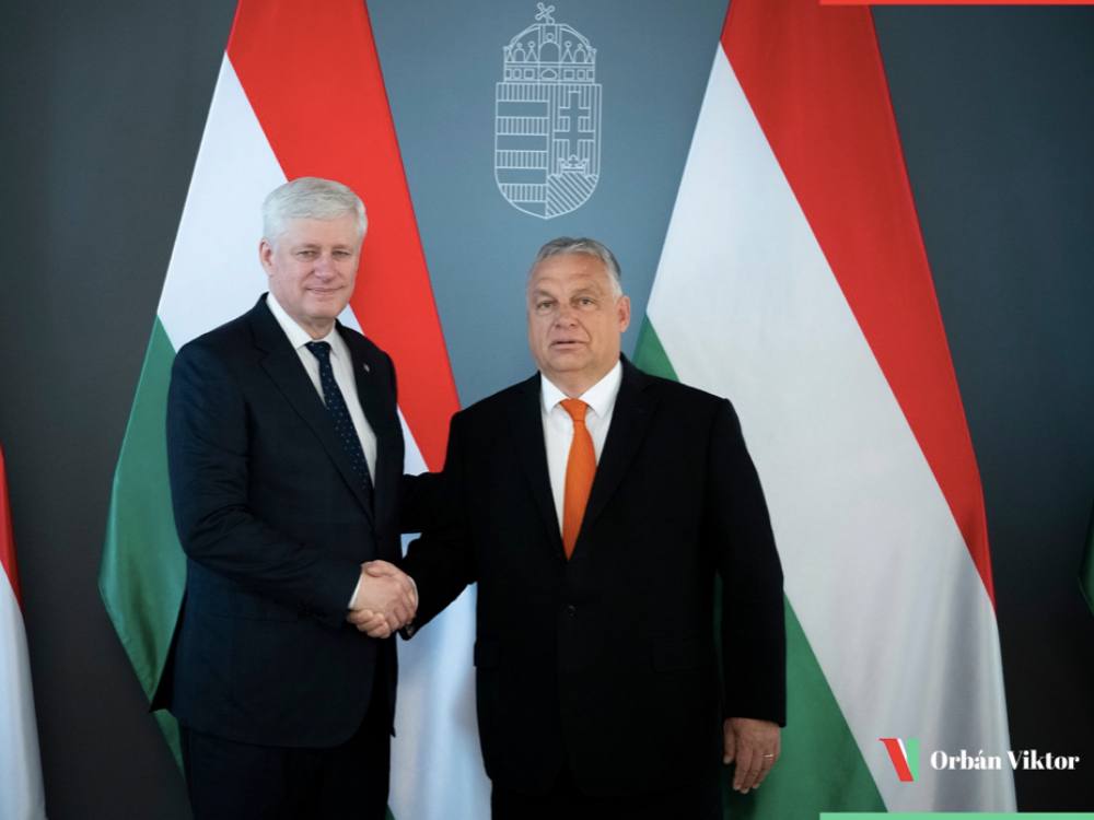 Stephen Harper shakes hands with Viktor Orbán in front of Hungarian flags. Both men wear dark suits and white shirts.