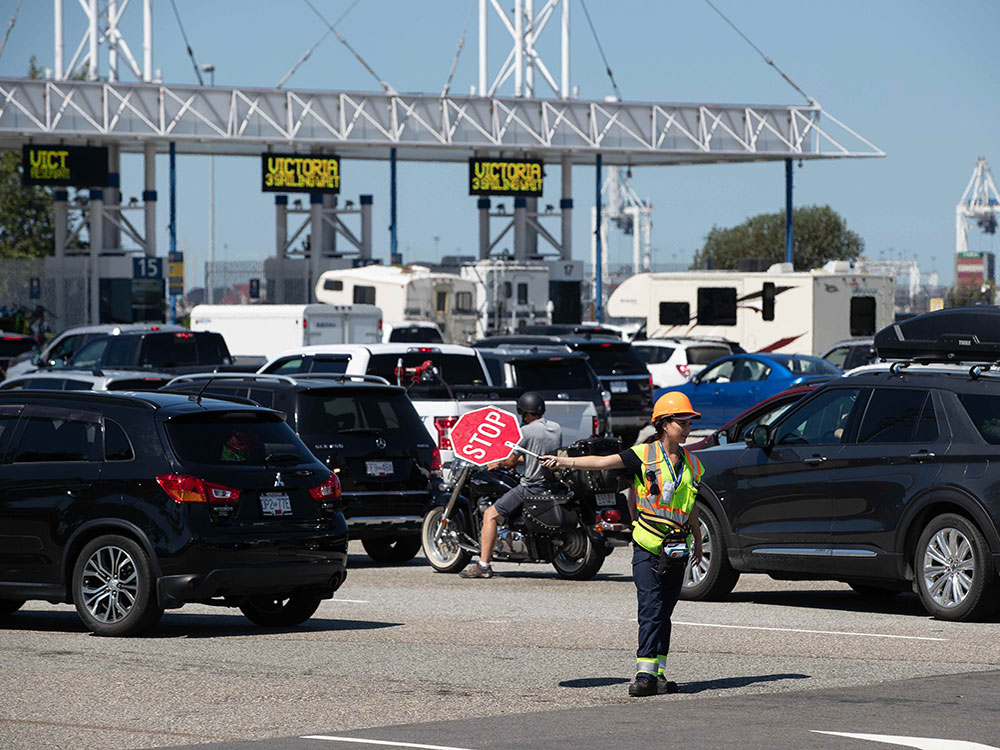 Vehicles are lined up outside the entrance to the Tsawwassen ferry terminal as a person in a safety vest and yellow hardhat holds out a stop sign.