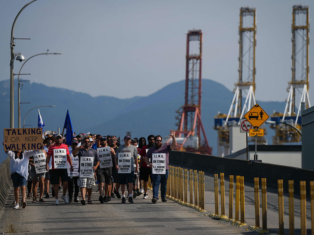 A large number of people carrying protest signs walk across a bridge toward the camera, with the Port of Vancouver’s giant cranes in the background.