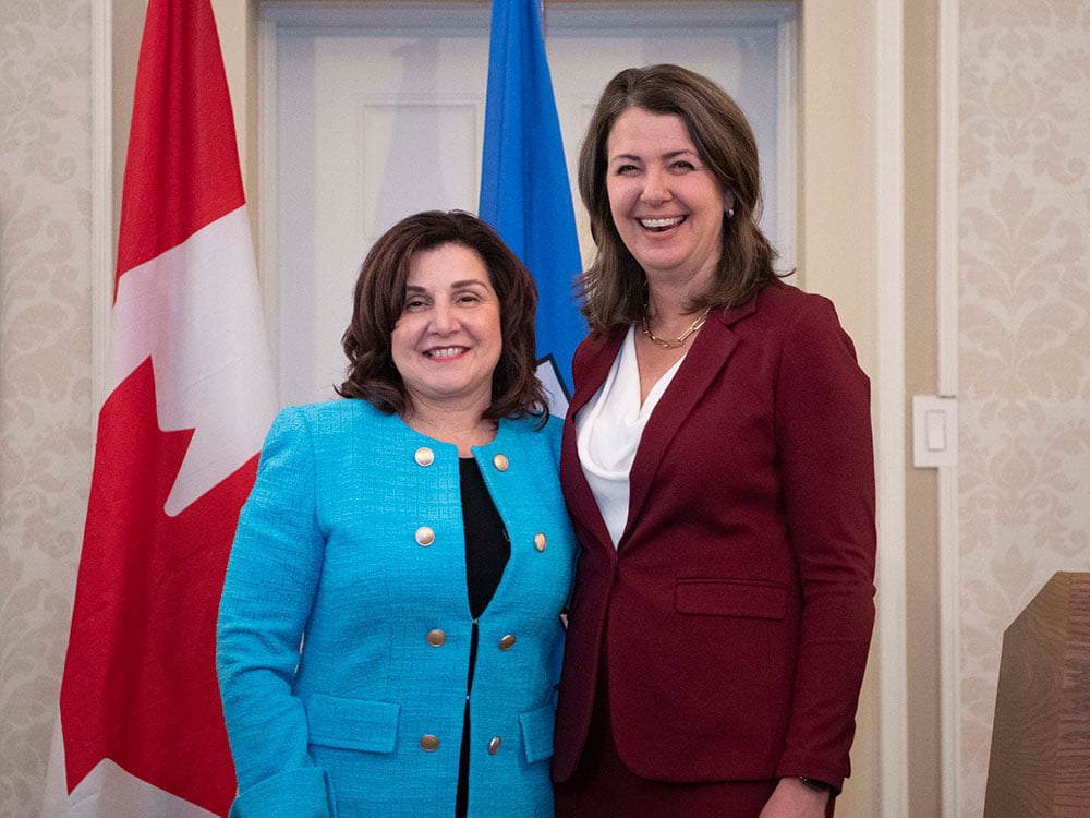 Adriana LaGrange, in a black dress and bright blue jacket, stands arm-in-arm with Danielle Smith. Canadian and Alberta flags are in the background.