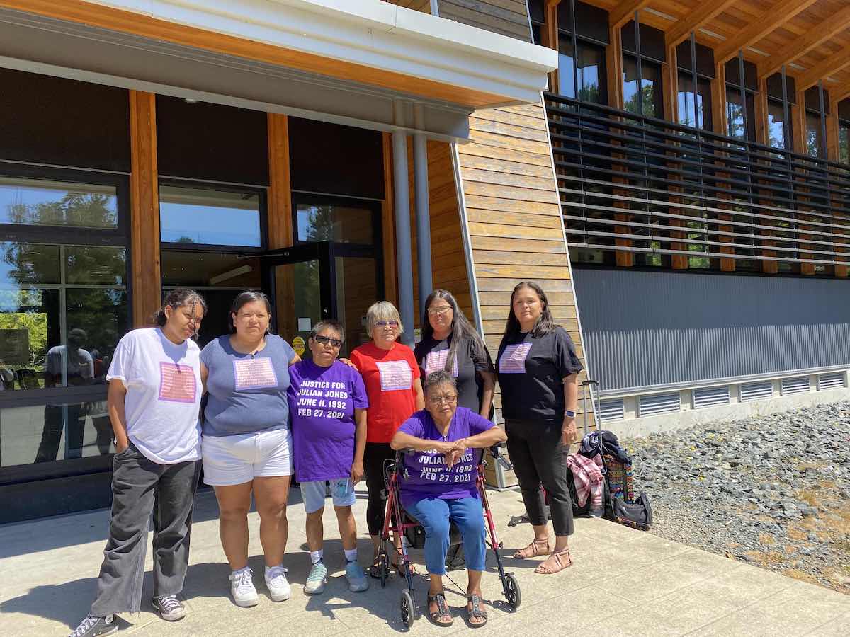 A group of Indigenous people stands in front of the Ty-Histanis Health Centre, a contemporary building with wood panelling. The group is wearing colourful T-shirts in memory of their relative Julian Jones.
