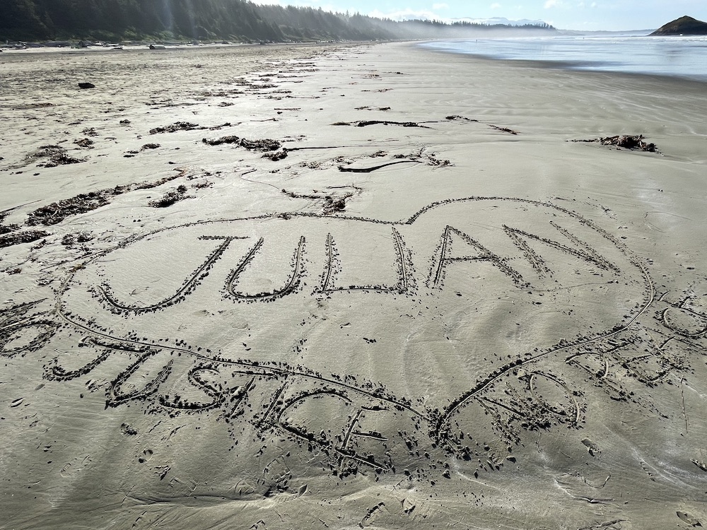 “Julian” is written in the sand inside a hand-drawn heart, under which are the words “No justice, no peace.” In the background, a winding stand of trees is to the left of the frame with surf on the sand to the right.