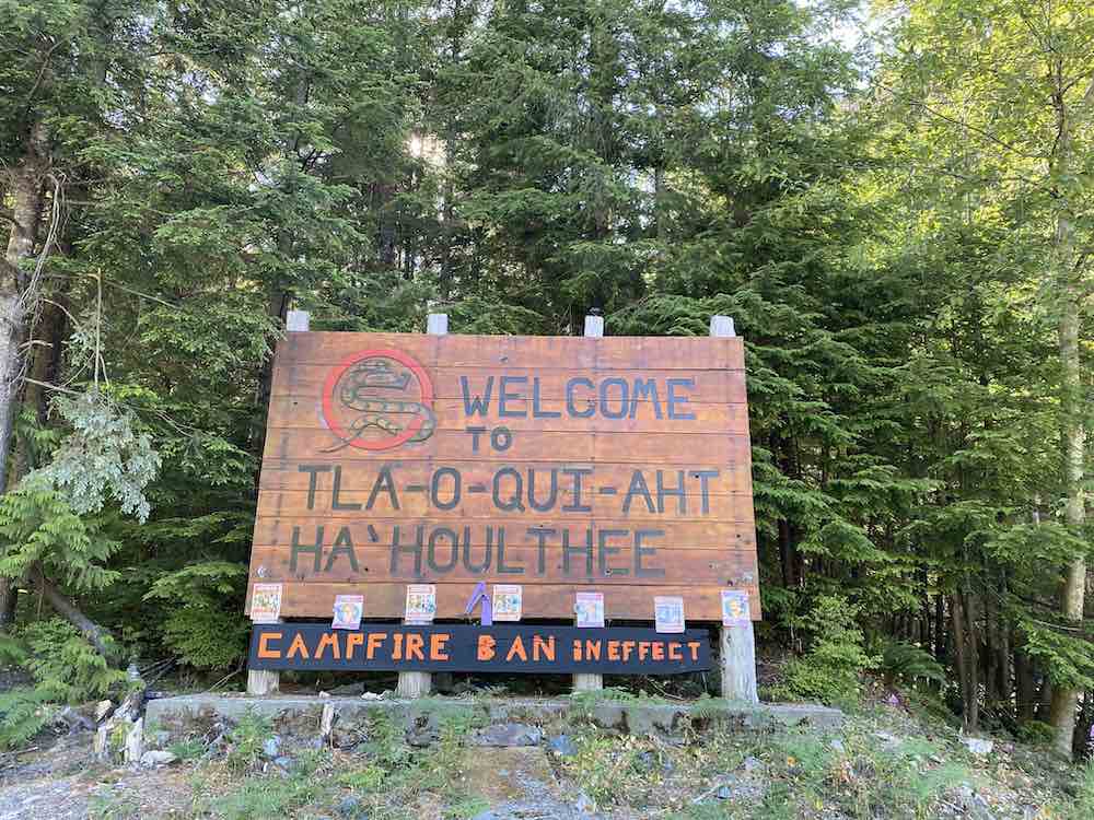 A large wooden welcome sign with black, all-caps text that reads “Welcome to Tla-o-qui-aht Ha’houlthee.” The sign also notifies visitors that a campfire ban is in effect.