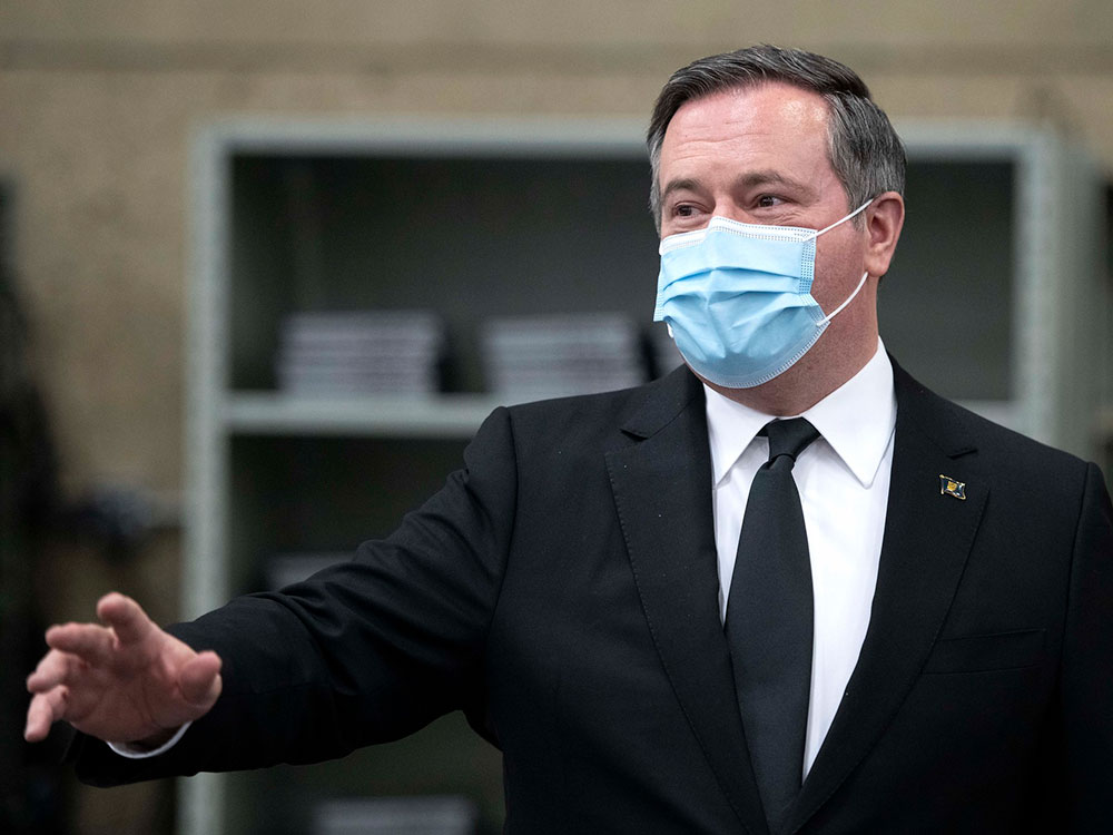 Jason Kenney, in a dark suit and tie, gestures behind a podium. He is wearing a pale blue mask.