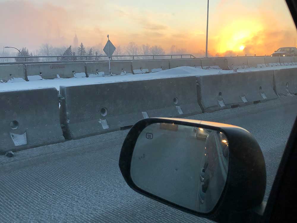 The view from the driver’s seat of a car at sunrise on a winter day. Concrete barricades are covered in a light dusting of snow. On the horizon, the grey sky is turning orange.