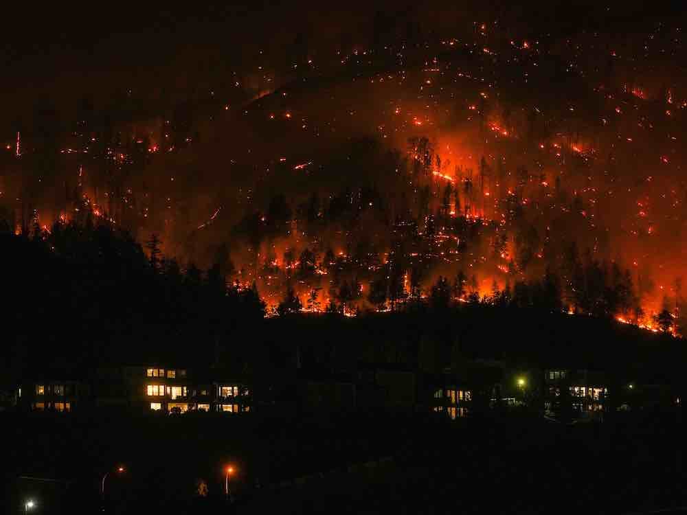 A hillside is covered in fire. In the foreground, several homes are visible in the night.