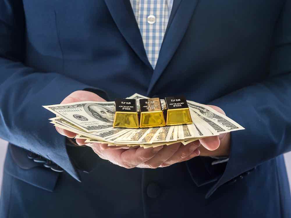 Close-cropped image shows the hands of a man in a suit holding gold bars and U.S. bills.