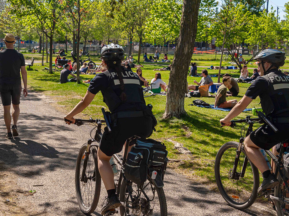 Two police officers in dark cycling uniforms ride their bikes along a walking path in a sunny park where people are walking and gathered in small groups on the grass. It’s a sunny day.