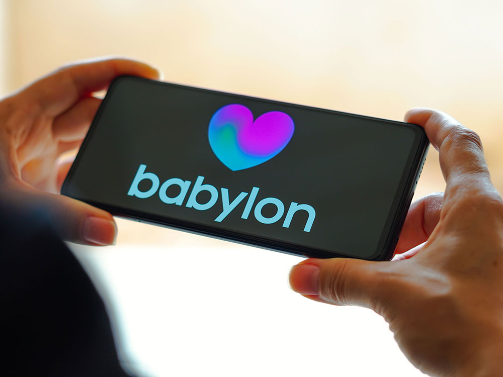 Hands hold a cellphone with Babylon’s logo — a blue and purple heart and the company name — on a black screen.