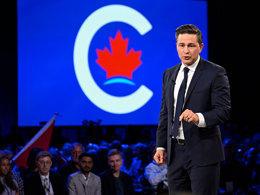 Pierre Poilievre, in a white shirt and blue suit and tie speaks in front of an audience. Behind him is a large Conservative party logo.