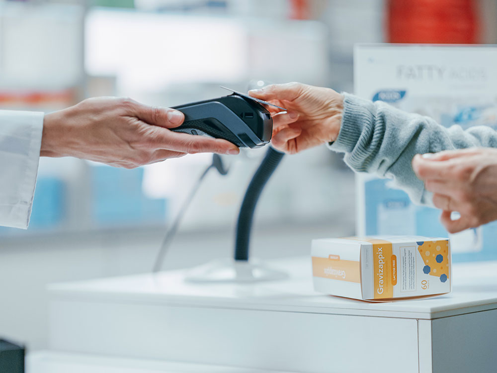 Two peoples’ hands reach across a white counter at a pharmacy. On the right, a pair of hands clad in grey knit sleeves holds a card over a black card reading payment system held by a hand clad in a white lab coat on the left. Under those arms sits a box of medication packaged in yellow, grey and white colourways. The light coloured background is in soft focus.