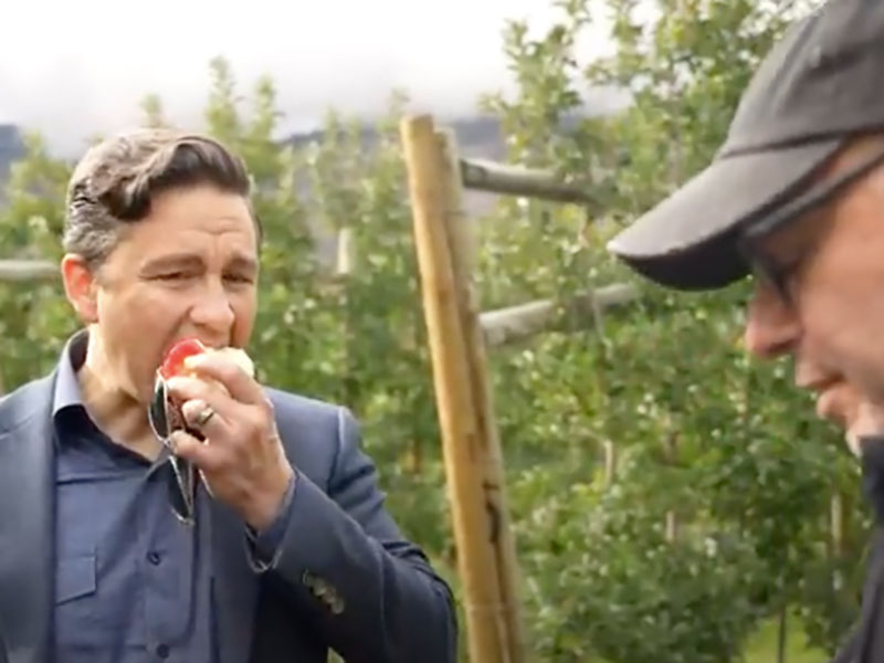 A white man in a navy suit takes a bite out of an apple while standing in an orchard.