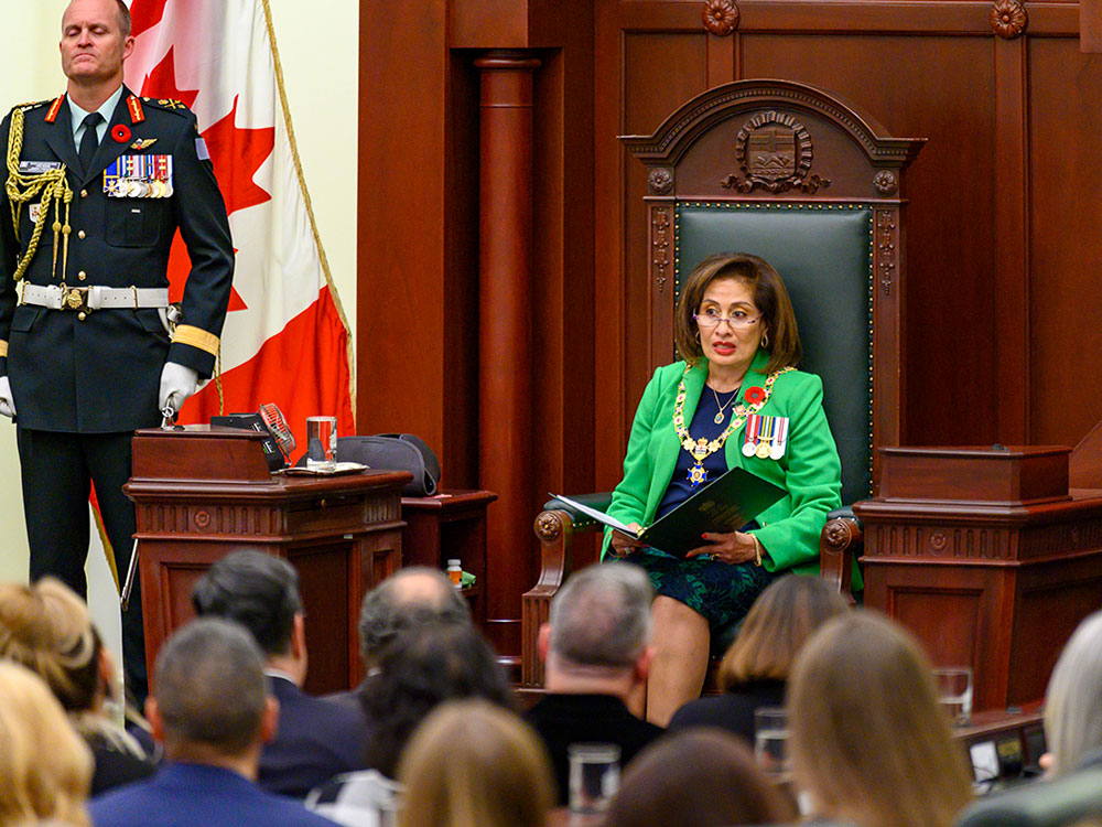 A woman with shoulder length brown hair and glasses sits in a large wooden chair with green leather padding. She wears a green jacker and blue shirt, a ceremonial chain of office and three medals, and holds a large binder.