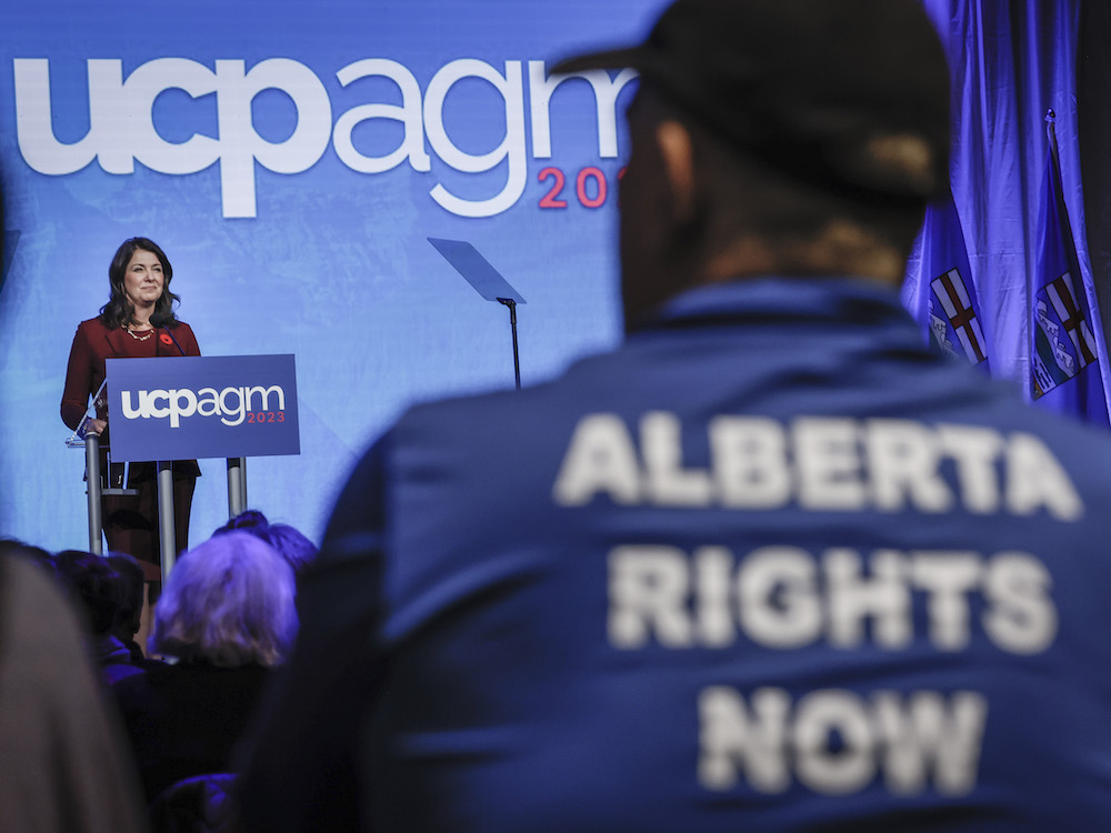 A white woman with long brown hair wearing a red suit and shirt stands at a podium. In the foreground, among the audience watching, is a man wearing a blue jacket with large white letters on the back saying "Alberta Rights Now."