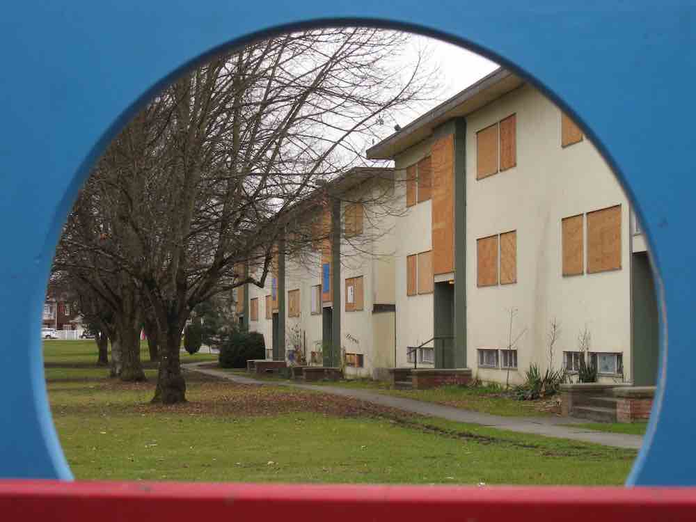 Framed by a blue circular border part of a children’s play structure, a row of beige three-storey walk-up apartment buildings stands along a tree-lined street. Their windows are boarded up with plywood.