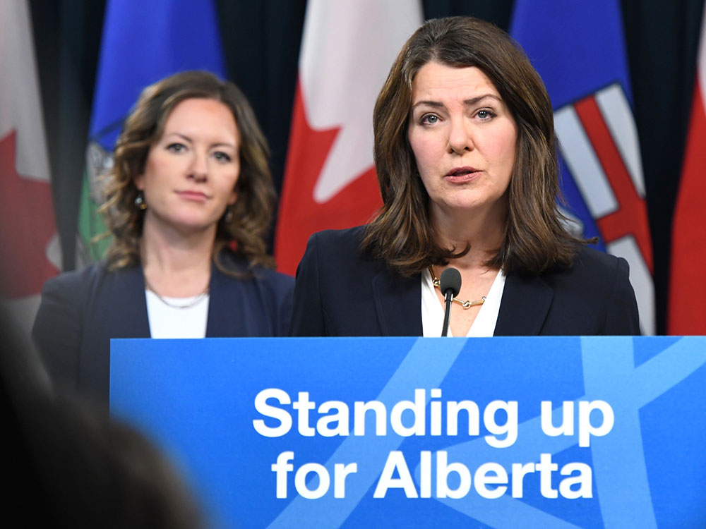 A white woman stands by a podium with a blue sign that says “Standing up for Alberta,” in front of Albertan and Canadian flags. She has shoulder length brown hair and wears a white shirt and blu jacker. Beside her is another white woman wearing similar clothes and also with shoulder length brown hair.