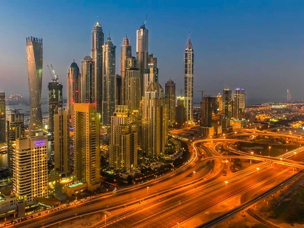 The skyline of Dubai at sunset, with a cluster of skyscrapers and a large tangle of freeways in the foreground.