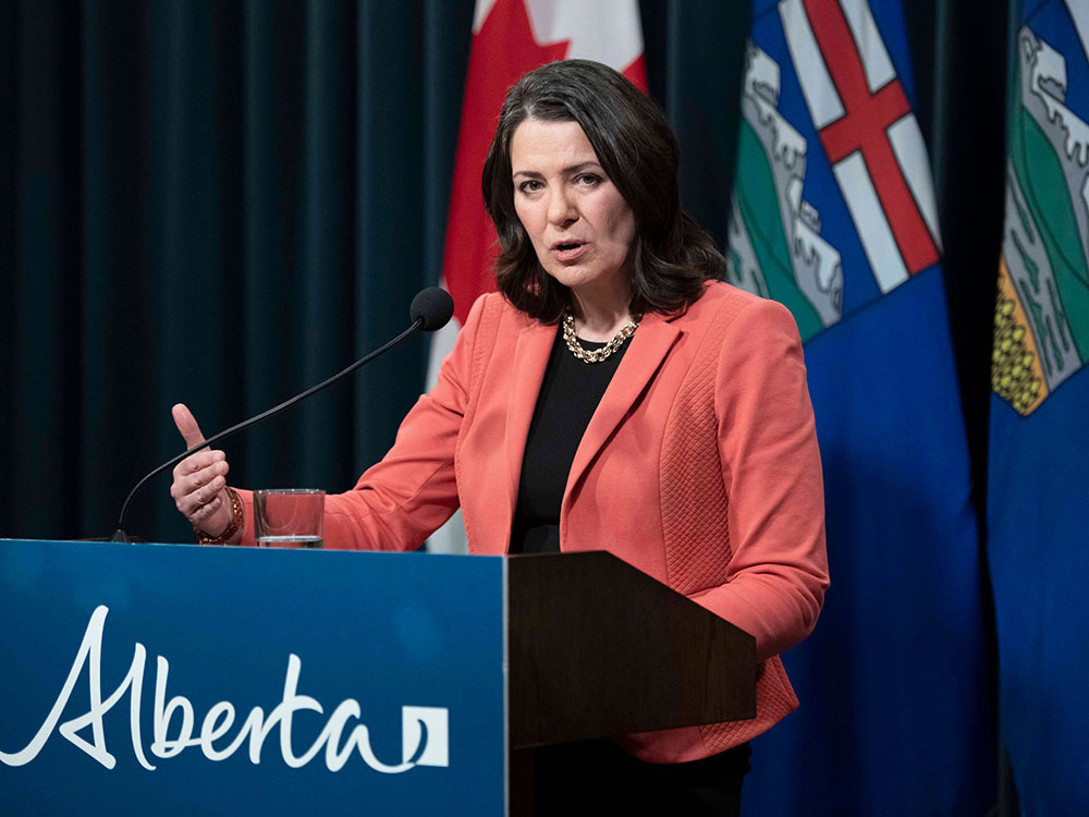 A 52-year-old white woman with shoulder-length dark hair stands at a podium. She wears a black shirt and rose blazer and stands in front of Canada and Alberta flags.