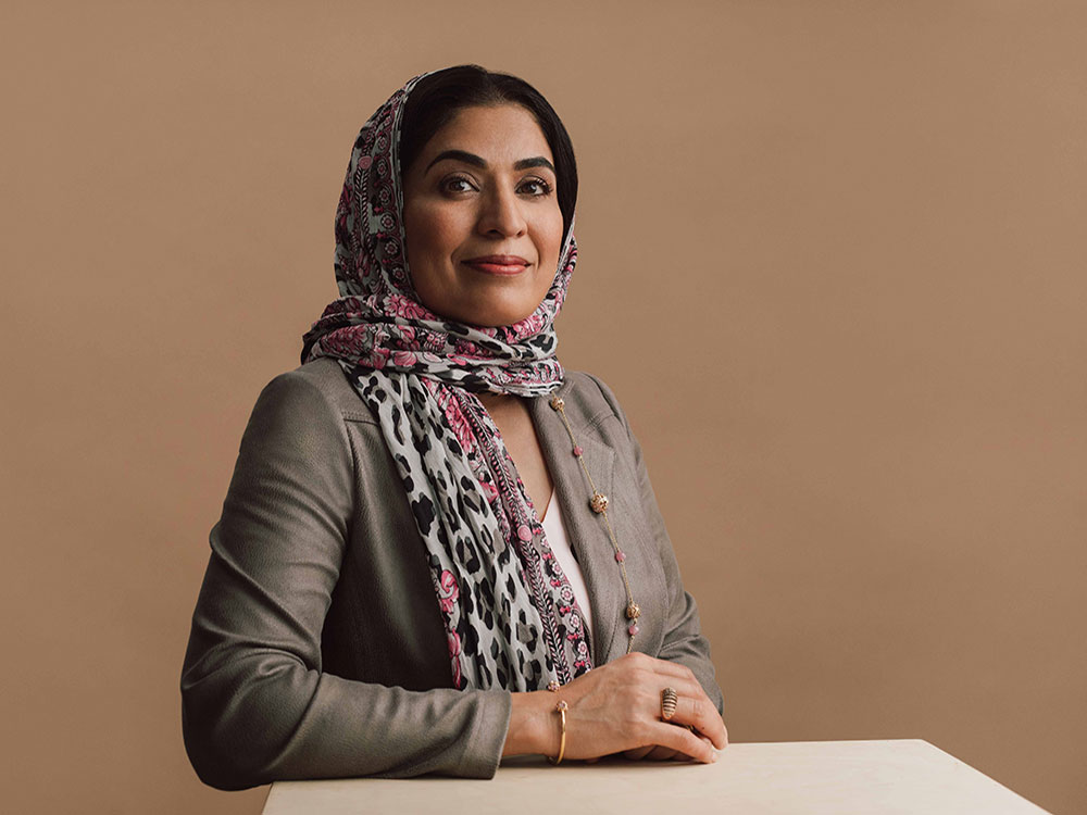 Fozia Alvi is seated against a beige studio background. She poses with her hand across a flat white surface and is looking at the camera directly, smiling slightly. She is wearing a headscarf with fuchsia floral and grey leopard print patterns, and a grey blazer. She has dark hair and brown eyes.