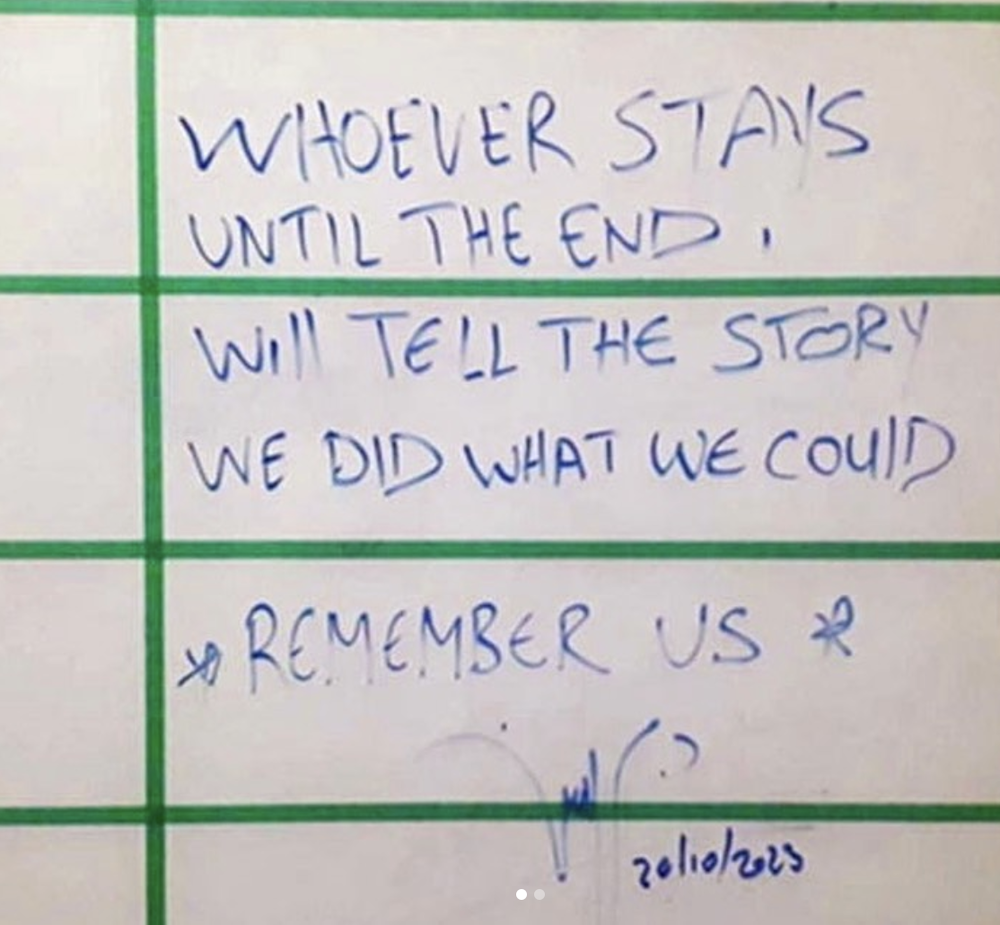 On a whiteboard with a wide grid of green masking tape reads a handwritten message using blue marker in all caps letters: 'Whoever stays until the end will tell the story. We did what we could. Remember us.'