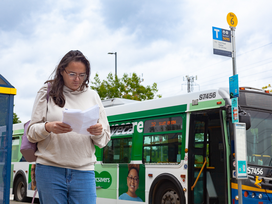 A young woman stands to the left of the frame in a cream-coloured hoodie, jeans and glasses. She holds a white piece of paper in front of her and is looking down, reading it. She is standing at a bus loop. Behind her, a white and green bus is parked with its doors open next to its blue stop.