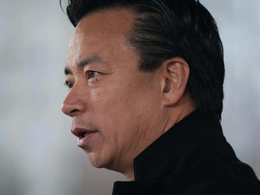 A 53-year-old man of Chinese heritage is captured in close-up against a grey background. He has slicked back black hair and wears a black jacket with the collar turned up.