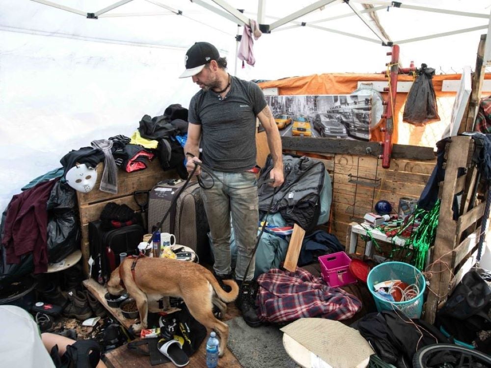 A light-skinned man with a dark hat, grey shirt and jeans holds the leash of a dog with brown fur. They are both standing underneath a tent, which provides shelter to belongings such as suitcases and clothes.