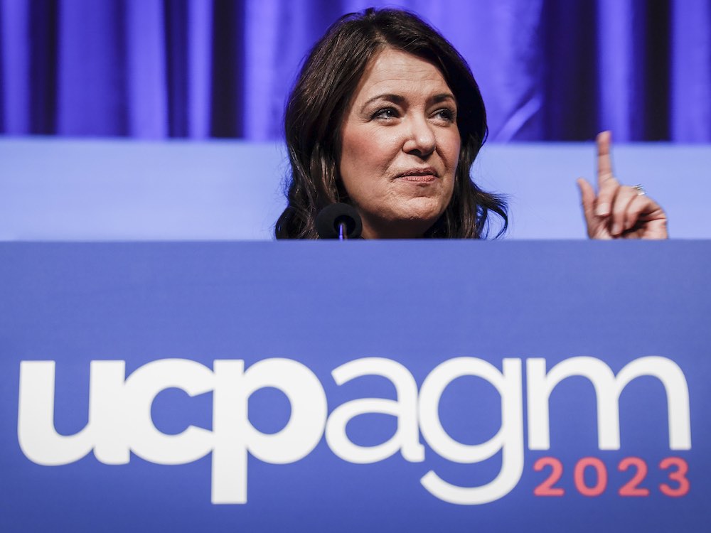 A 52-year-old white woman with long dark hair stands behind a podium with a large blue sign that says 'ucpagm 2023.' She gestures, pointing skyward with one index finger.