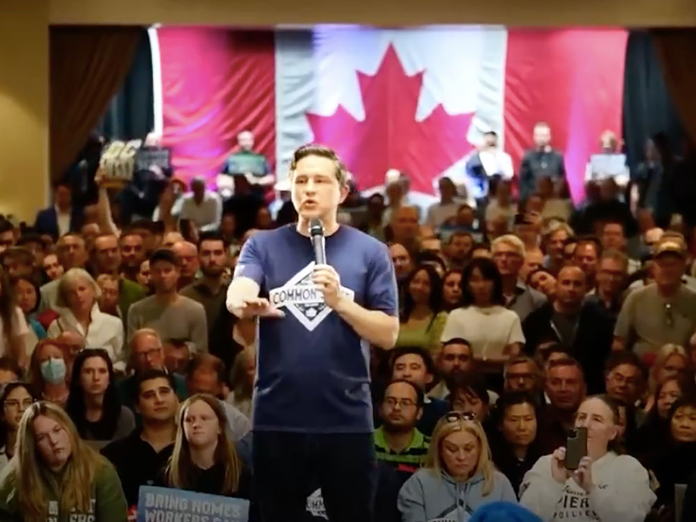 A 44-year-old white man with short dark hair stands in front of an audience and a Canadian flag, holding a microphone. He’s wearing a blue T-shirt and black jeans.