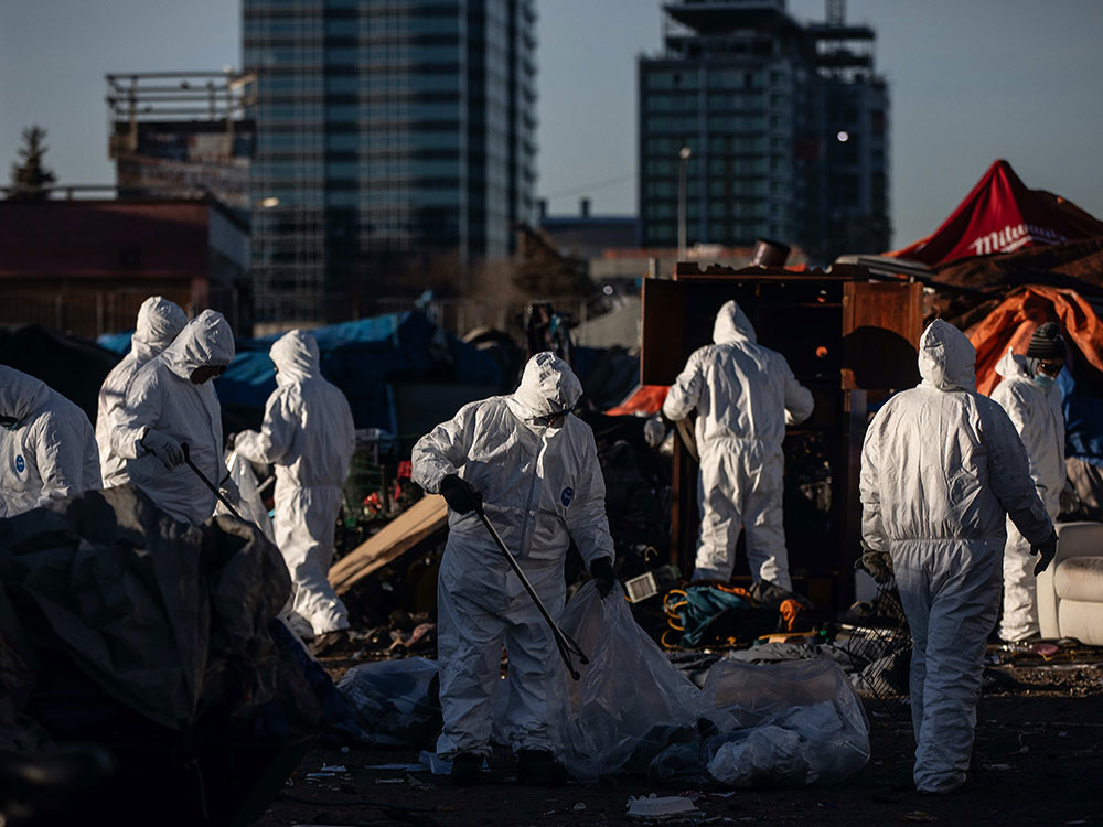 Eight people in white biohazard suits stand in front of tents. Some are loading people’s possessions into trash bags.
