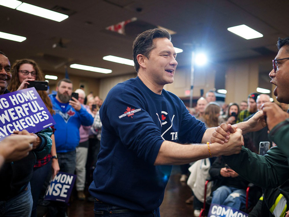 Pierre Poilievre has wavy brown hair and light skin. He is in the middle of the frame in profile. He is smiling enthusiastically while shaking hands with a man with glasses and dark skin. They are surrounded by supporters in an indoor reception area. People are smiling, cheering and holding placards with white sans-serif text over a navy background. One reads 'Bring home powerful paycheques.'
