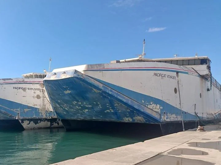 Two catamarans, looking neglected, at dock. They have a blue lower hull with white above.