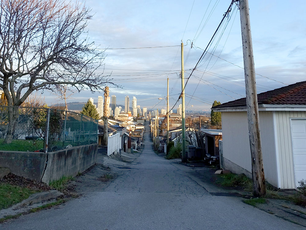 An alleyway runs through East Vancouver facing a skyline of highrises. A telephone pole reaches into a blue sky.