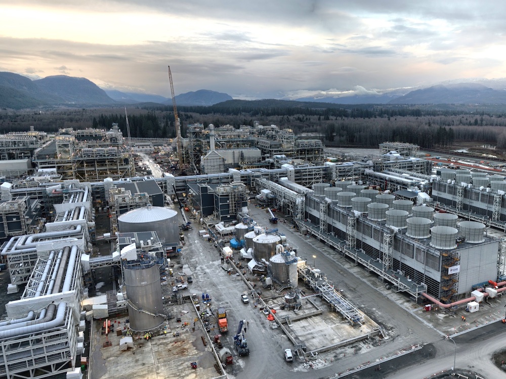 The photo shows a giant LNG plant with rows of metal structures and pipes sprawling across a large site. In the background are forests and mountains.