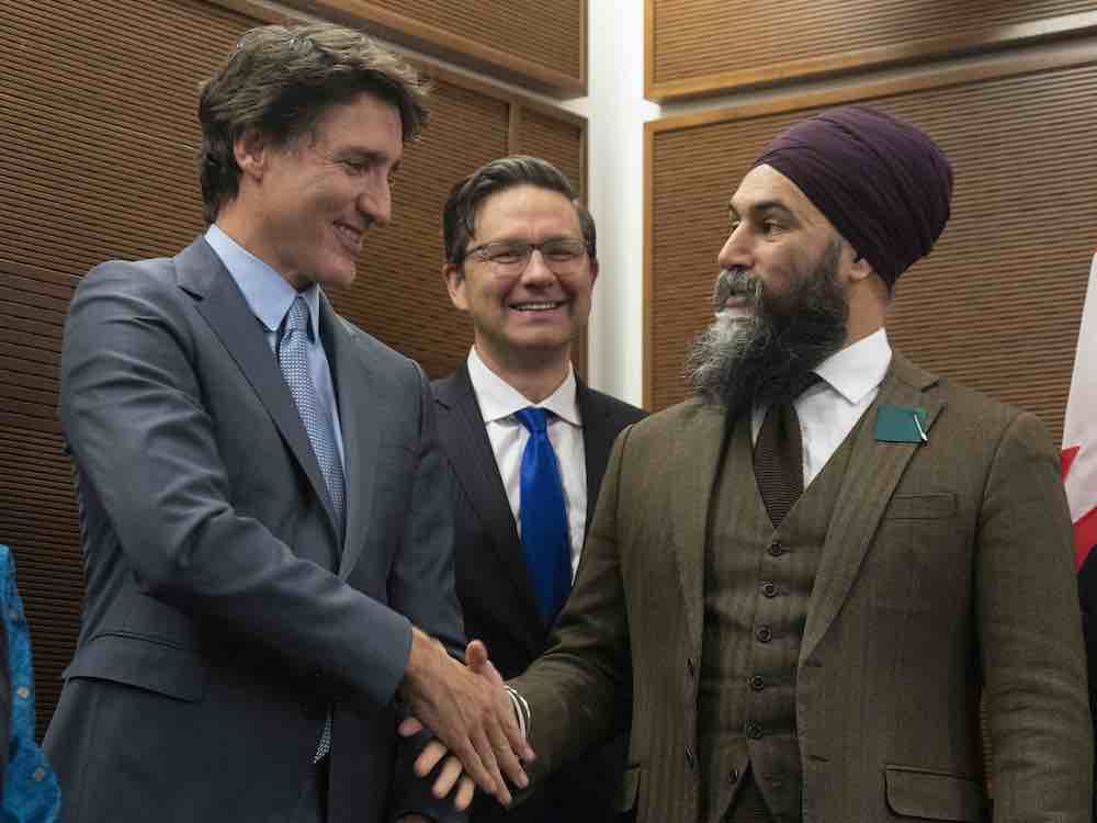 Three men in suits, one wearing a turban, pose for the camera. Two are shaking hands; the third looks on from behind, smiling.