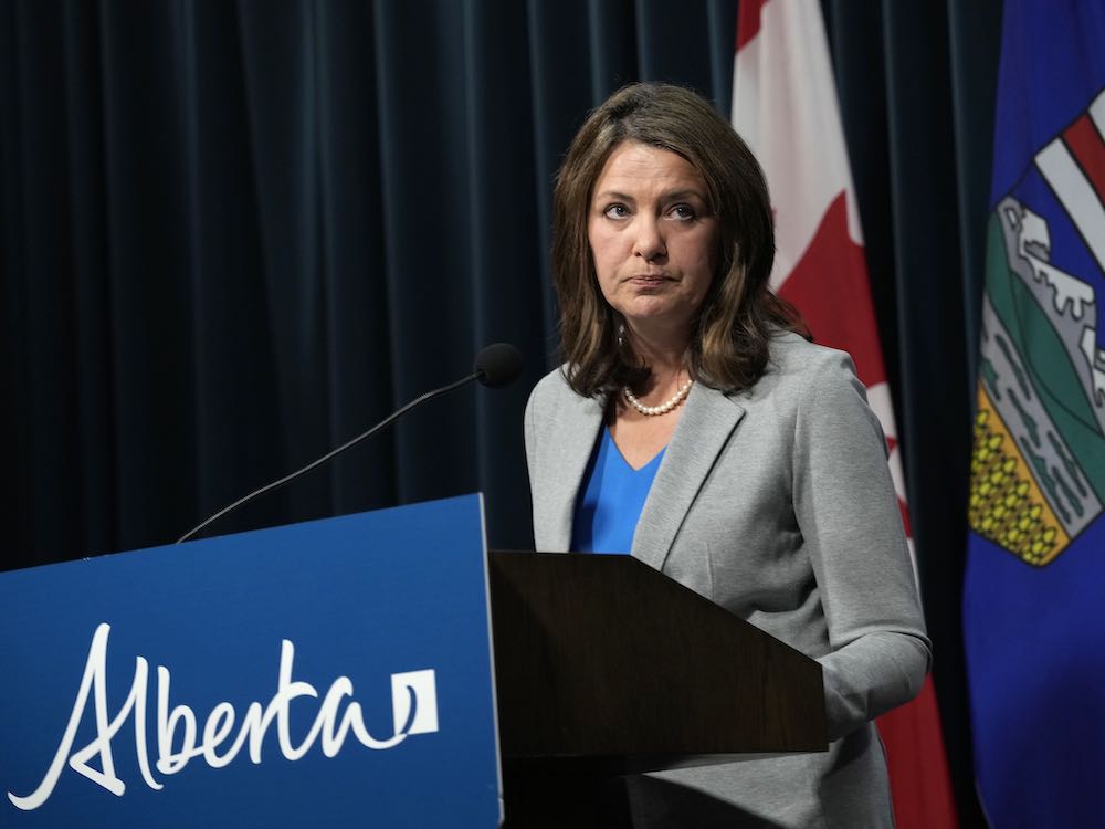 Danielle Smith is a white woman with shoulder-length brown hair. She wears a grey blazer over a bright blue top and looks up towards the right of the frame into the middle distance. She stands at a podium with a blue 'Alberta' sign affixed to it. Behind her are the Canadian and Alberta flags against a row of blue curtains.
