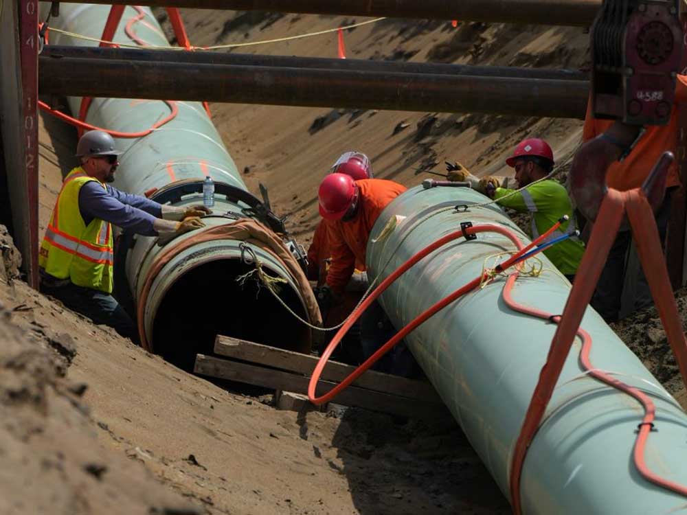 Two very large sections of pale green pipe are in a large, sandy trench, with workers in safety gear apparently trying to connect them.