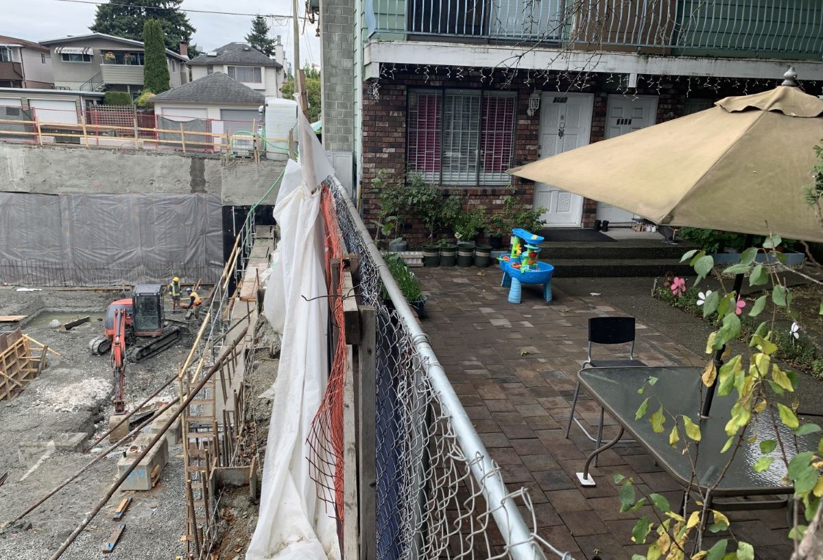 On the left side of the image is a crew working on the foundations of a new development beside an orange digging machine. On the right side, separated by a metal fence, is the patio and garden of a multi-unit home with a balcony above it. There is a blue children's toy on the patio. There is also a glass table, metal chairs and an umbrella.