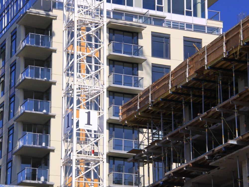 A multi-storey residential building is under construction in Vancouver. An exterior elevator rises up the side of the building. In the right foreground is another unfinished building.