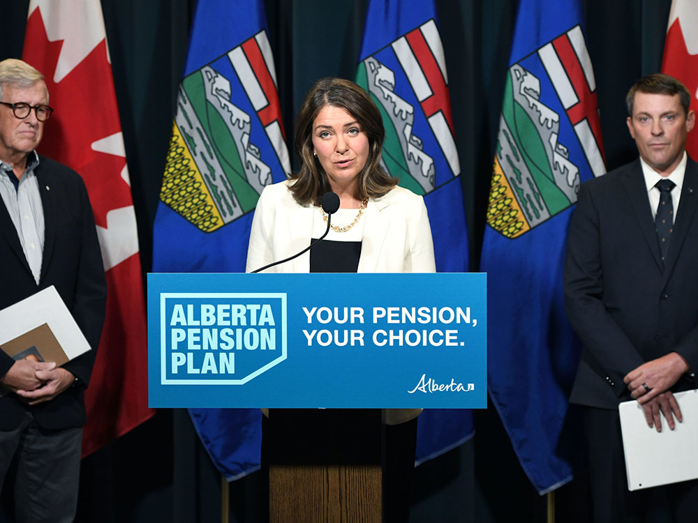 A silver-haired light-skinned person in a blue blazer and white shirt stands to the left of a middle-aged woman with shoulder length dark hair and a white jacket, black shirt and gold chain. To her right is a younger man in dark suit and tie and a white shirt. Alberta and BC flags are behind them. the woman stands at a podium with a sign that says “Alberta Pension” and “Your Pension, Your Choice.”