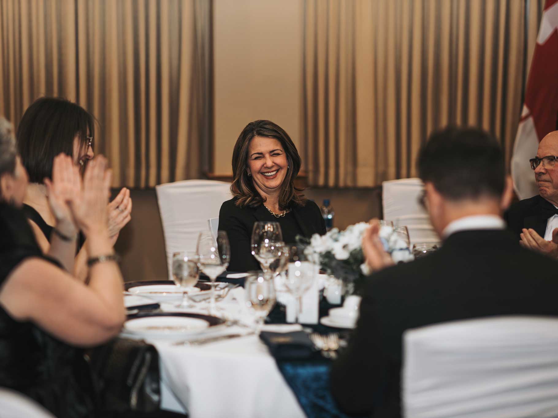A 50-ish woman with shoulder-length dark hair and a dark suit smiles at the centre of the photo, with others applauding her round a fancy dinner table.