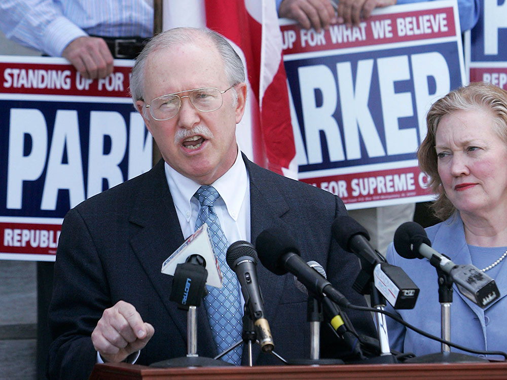 A grey-haired, light-skinned older man in a suit and shirt tie, with a grey moustache speaks in front of a background of campaign signs.