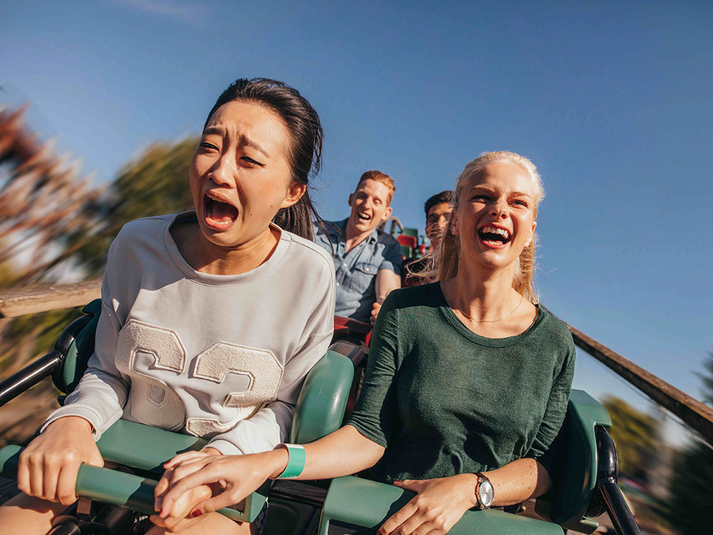 Two young people are on a roller-coaster facing the camera, holding hands. The one on the right is smiling, while the other looks terrified.