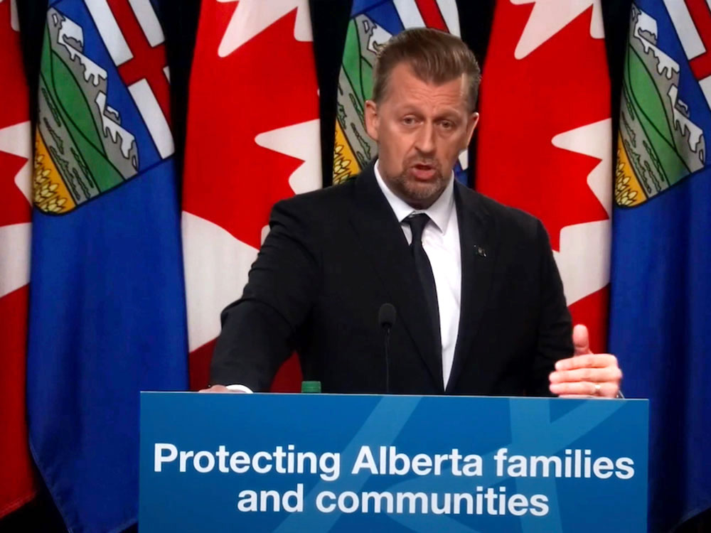 A middle-aged light-skinned man stands at a podium with a sign that says 'Protecting Alberta families and communities.' He wears a black suit, white shirt and black tie.