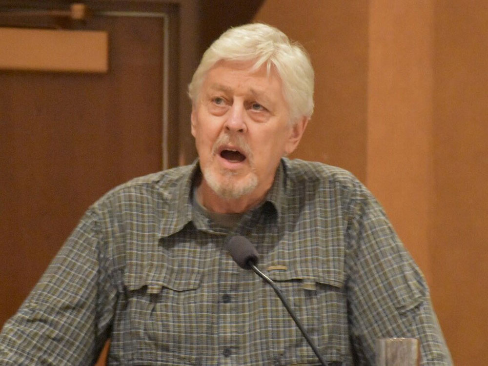 A lighter-skinned man with white hair and goatee, wearing a checkered, collared shirt, speaks at a podium before a small microphone.