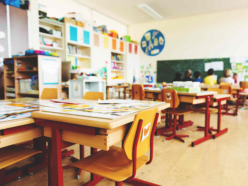The interior of an elementary school classroom features wooden desks and chairs grouped together in pods and colourful paper stuck on white shelves. In the far background in soft focus is a group of students seated at a table.