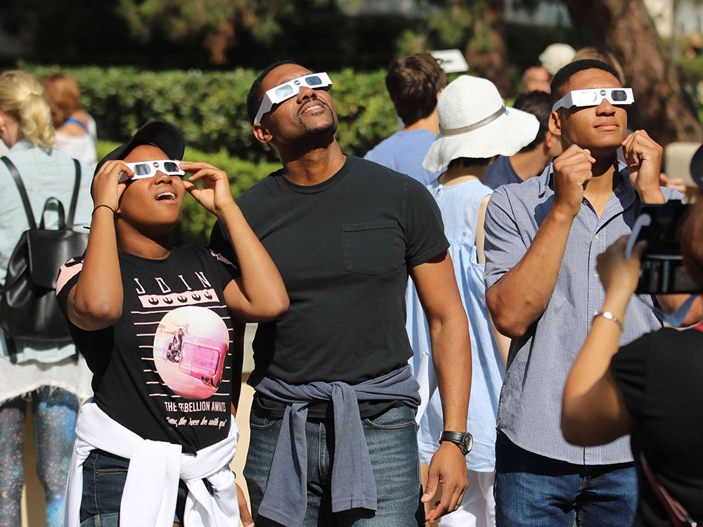 A crowd of people stand together outdoors, wearing paper sunglasses and looking up at a solar eclipse. In the foreground is a couple with dark skin wearing black T-shirts, looking up in awe.