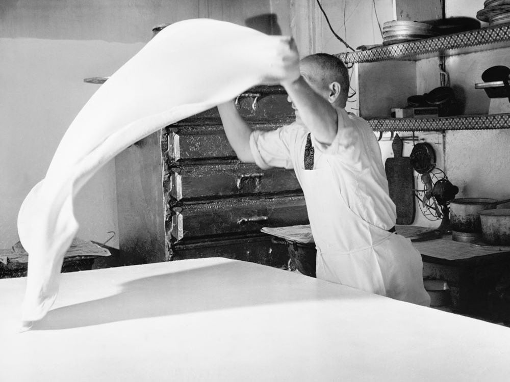 A black and white photo of a man who is making pizza dough by tossing it. 
